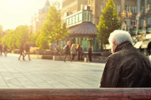 view from behind of retired senior man sitting on a bench in a city with people walking by in the background